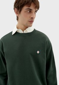 Felpa verde con scollo rotondo e un patch testurizzato che presenta un motivo a raggi di sole. Il colletto è bianco e con bottoni, offrendo un contrasto.