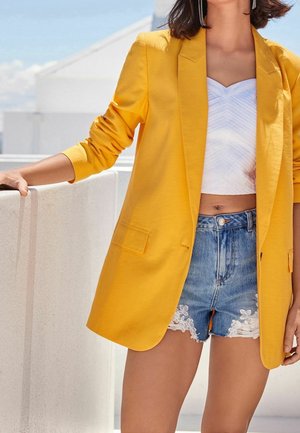 Keltainen bleiseri pitkillä hihoilla, yhdistettynä valkoiseen lyhytlahkeiseen toppiin, jossa on aaltoilukuosi, sekä denimshortseihin, joissa on pitsisomisteet.