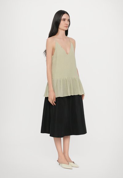 Light green, pleated blouse with a v-neck and tiered hem, paired with a black, flowy skirt. Model wears off-white pointed shoes.