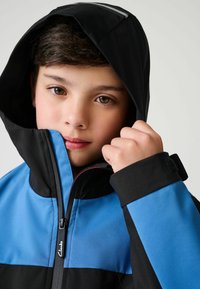 Schwarze und blaue Jacke mit durchgehendem Reißverschluss, Kapuze und verstellbaren Bündchen mit strukturierter Oberfläche. Logo-Detail befindet sich am Reißverschluss.