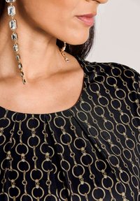 Frau trägt eine schwarze Bluse mit beigem Ringmuster und langen rechteckigen Kristallohrringen.