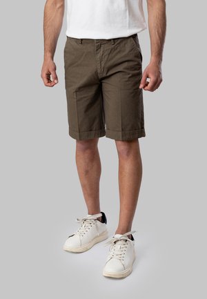 Uomo che indossa pantaloncini verdi oliva fino al ginocchio, sneakers bianche con dettagli neri e una maglietta bianca, in piedi su uno sfondo grigio.