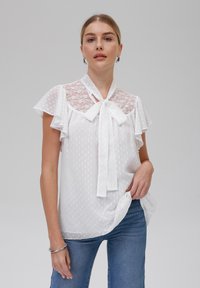 Hvit bluse med blondedetalj og prikkete mønster. Har volangede ermer og en halsknute. Kombinert med blå jeans.