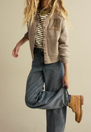  béžový pletený cardigan s písmenem "A" na přední straně, pod ním pruhované zelenobílé tričko, volně padnoucí modré džíny a hnědé šněrovací boty.