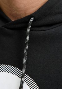 Sort hættejakke med en struktureret stof. Har en sort og hvid stribet snor og et cirkulært grafisk design på brystet.