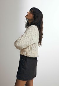 Chunky strikket cremesweater med kabelmønstre og frynsekant, parret med en mørk, struktureret miniskørt. Ensfarvet baggrund.