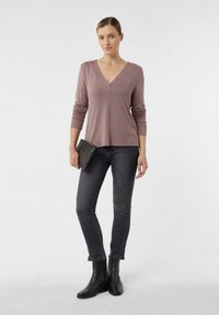 Graues Langarmoberteil mit tiefem V-Ausschnitt, kombiniert mit dunkelblauen Jeans und schwarzen Knöchelstiefeln. Hält eine strukturiertes schwarzes Clutch.