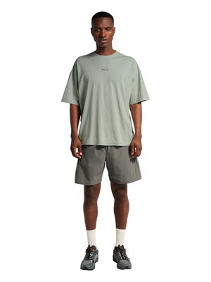 Mężczyzna stoi, ubrany w oversize'ową koszulkę w kolorze szałwii z napisem "HALO", szare szorty, białe skarpetki i szare trampki, zwrócony przodem.