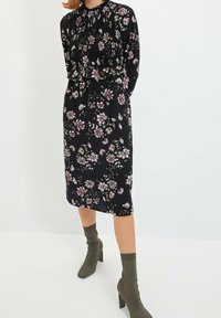 Kobieta nosi czarną, kwiecistą sukienkę do kolan z długimi rękawami i marszczonym bodicem, w połączeniu z oliwkowymi, z wysokim obcasem, skarpetkowymi botkami.