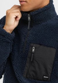 Navy fleece pullover features a half-zip collar, textured material, and a contrasting black zippered chest pocket with logo detail.