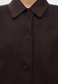 Gros plan d'un manteau en laine brun foncé avec un col classique et des boutons avant dissimulés porté par une personne.