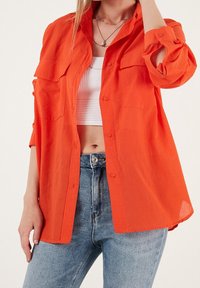 Femme portant un jean bleu clair, un débardeur côtelé blanc coupé et une chemise orange vif ample avec les manches retroussées.