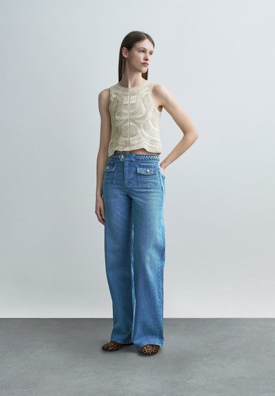 Νεαρή γυναίκα στέκεται με το χέρι στη μέση, φορόντας κρεμ πλεκτό αμάνικο τοπ, μπλε τζιν παντελόνι φαρδιές παντελόνι με μπροστινές τσέπες και παπούτσια με λεοπάρ μοτίβο.