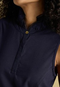 Marinefarbener ärmelloser Oberteil mit Stehkragen, ausgestattet mit einer Knopfschließe und aufwendigen Stickdetails entlang des Ausschnitts. Weicher, glatter Stoff.