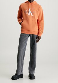 Orange hooded sweatshirt with front pocket and white "Calvin Klein Jeans" logo, paired with gray wide-leg denim trousers and black footwear.