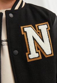 Svart varsityjacka med en stor, texturerad vit "N"-applikation omgiven av brunt. Har svarta läders ärmar och en randig krage.