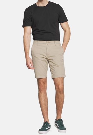 Mann in einem schwarzen T-Shirt, beigen Knie-langen Shorts mit Taschen und grünen Sneakers, der vor einem schlichten hellen Hintergrund steht.