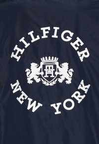Granatowa tkanina z białą haftowaną dekoracją. Zawiera napisy "HILFIGER" i "NEW YORK", otoczone dwoma motywami lwów oraz herbem z koroną pośrodku.