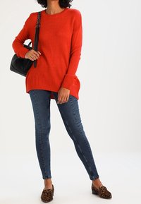 Červený pletený svetr s kulatým výstřihem, kombinovaný s těsnými modrými skinny džíny a slip-on botami s leopardím vzorem. Černá kabelka s řetízkovým popruhem.