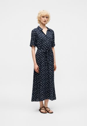 Femme aux cheveux blonds bouclés portant une robe midi bleu marine à manches courtes, boutonnée, imprimée de fleurs blanches et ceinturée, debout en sandales marron.