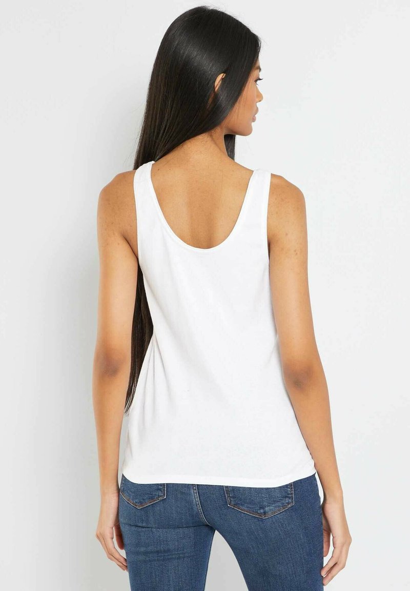 Kobieta z długimi ciemnymi włosami, ubrana w białą bezrękawnik i niebieskie dżinsy, widziana z tyłu na jednolitym jasnym tle.