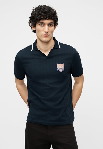 Νέος με σκούρα σγουρά μαλλιά, που φοράει μπλε κοντομάνικη πόλο μπλούζα με έμβλημα στο στήθος, στέκεται με τα χέρια πίσω από την πλάτη.