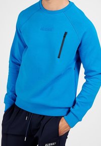 Sudadera azul con cuello redondo, puños acanalados y un bolsillo con cremallera en el lado izquierdo del pecho; fabricada con una tela suave, presenta un detalle de logo.