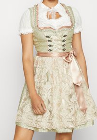 Robe dirndl verte et ivoire avec superposition en dentelle brodée, ceinture en ruban de satin, motif floral, et blouse blanche avec bordure en dentelle.
