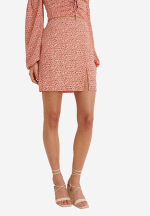 Mini-jupe à motif floral en corail, avec une fente sur le côté. Associée à des sandales à talons à lanières beiges, mettant en valeur une texture lisse.