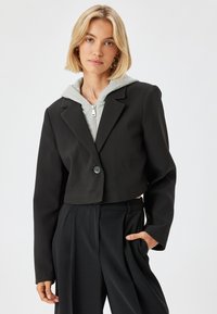 Blazer negro cortado con cierre de botón, que presenta un diseño a medida, mangas largas y una textura suave, superpuesto sobre una sudadera con cremallera gris.
