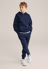 Tumed sinine kapuutsiga sviiter ja trennipüksid, mis on valmistatud pehmest kangast. Kapuutsil on ees tasku ja logo detail; pükstel on kitsenev lõige.