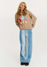 Jasnobrązowy sweter dzianinowy z kolorowymi aplikacjami grzybów; w zestawie z szerokimi dżinsami w jasnoniebieskim kolorze. Czarne buty na platformie.