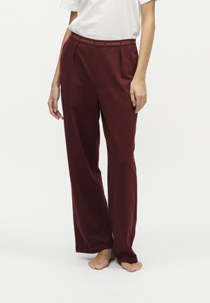 Femme portant un pantalon bordeaux à jambes larges avec une ceinture marquée Lacoste et un haut blanc à manches courtes, debout pieds nus sur fond blanc.