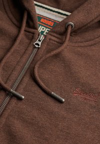 Sudadera con cremallera marrón, con cordones y puntas metálicas grabadas "SuperDry", y logo bordado "SuperDry" en el pecho, mostrando parcialmente la etiqueta interior de la marca.