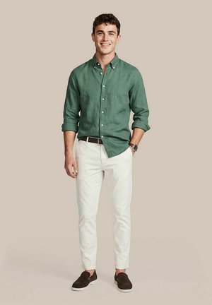 Jonge man staand, glimlachend, draagt een groen overhemd met knopen, witte broek, bruine riem, horloge en donkerbruine instapschoenen tegen een beige achtergrond.