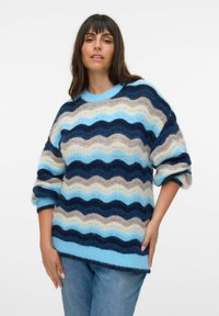 Trui in blauw, beige en marineblauwe zigzagpatroon; zachte textuur; ronde halslijn; losse pasvorm; driekwartmouwen. Gecombineerd met blauwe jeans.