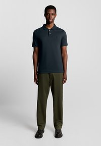 Mörkgrå kortärmad polo med krage, med en broderad logotyp på bröstet, tillsammans med olivgröna byxor och svarta skor.