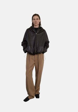 Chaqueta de cuero marrón oscuro con capucha y bolsillos delanteros, combinada con pantalones de pierna ancha beige. El modelo se encuentra de pie frente a un fondo blanco liso.