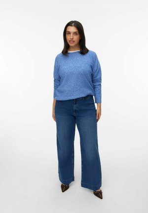 Pull en maille texturée bleu avec un col rond associé à un jean en denim bleu à jambes larges et des escarpins marron pointus avec des accents cloutés.