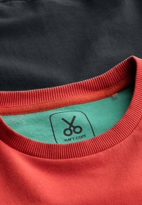 Felpa rossa in cotone con collo a coste, indossata sopra un indumento nero. L'etichetta interna verde presenta un logo a forbici e la scritta "KAFT.COM."