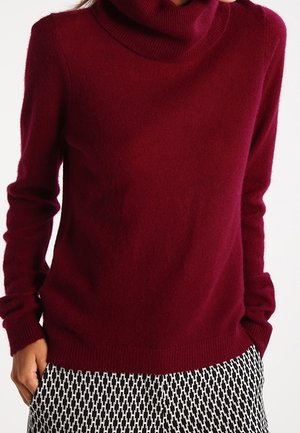 Vrouw draagt een bordeauxrode coltrui met lange mouwen, gecombineerd met zwart-wit geruite broek. Handen deels in de zakken.