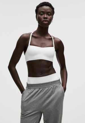 Vrouw met kort haar, gekleed in een witte sportbeha en grijze joggingbroek, met handen in de zakken, staand tegen een effen lichte achtergrond.