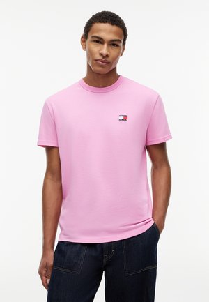Νέος άνδρας που στέκεται με το ένα χέρι στην τσέπη, φορώντας ένα ροζ κοντομάνικο T-shirt με μικρό λογότυπο στο στήθος και σκούρο τζιν.