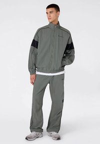 Trainingsjack en broek in gedempt groen met zwarte accenten. Voorzien van een ritssluiting aan de voorkant, elastische manchetten en een logovlek op het been.