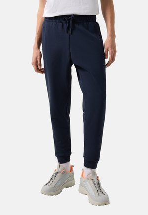 Muž oblečený v tmavě modrých teplákových kalhotách, bílých ponožkách, šedých a oranžových teniskách a zaťaté bílé tričko, stojící uvolněně se sepnutýma rukama.