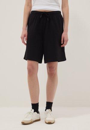 Persona vistiendo pantalones cortos negros con cordón, zapatillas blancas, calcetines negros y una camiseta blanca, de pie contra un fondo liso y claro.