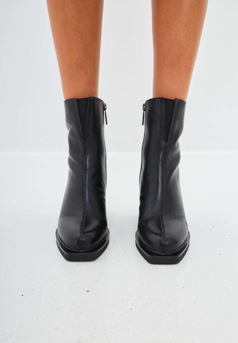 Botas negras hasta el tobillo de piel suave, con un diseño elegante, cremallera frontal, puntera cuadrada y tacón bajo apilado.