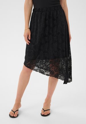 Jupe midi noire en dentelle avec motif cachemire et ourlet asymétrique, portée avec un top noir et des tongs noires sur un mannequin féminin.