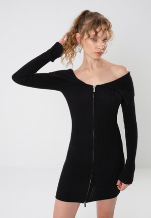 Sort ribbestrikket kjole med lange ermer, off-shoulder-design, glidelås foran og en kroppsformet silhuett.