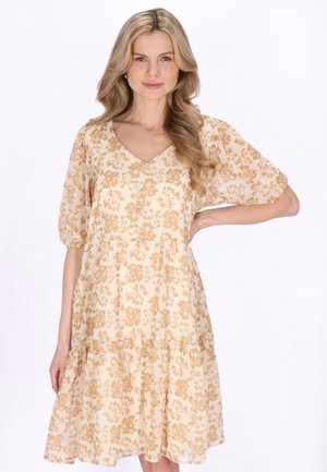 Kobieta o długich blond włosach, ubrana w beżową sukienkę w kwiaty sięgającą do kolan z bufkami na rękawach, stojąca z jedną ręką na biodrze.